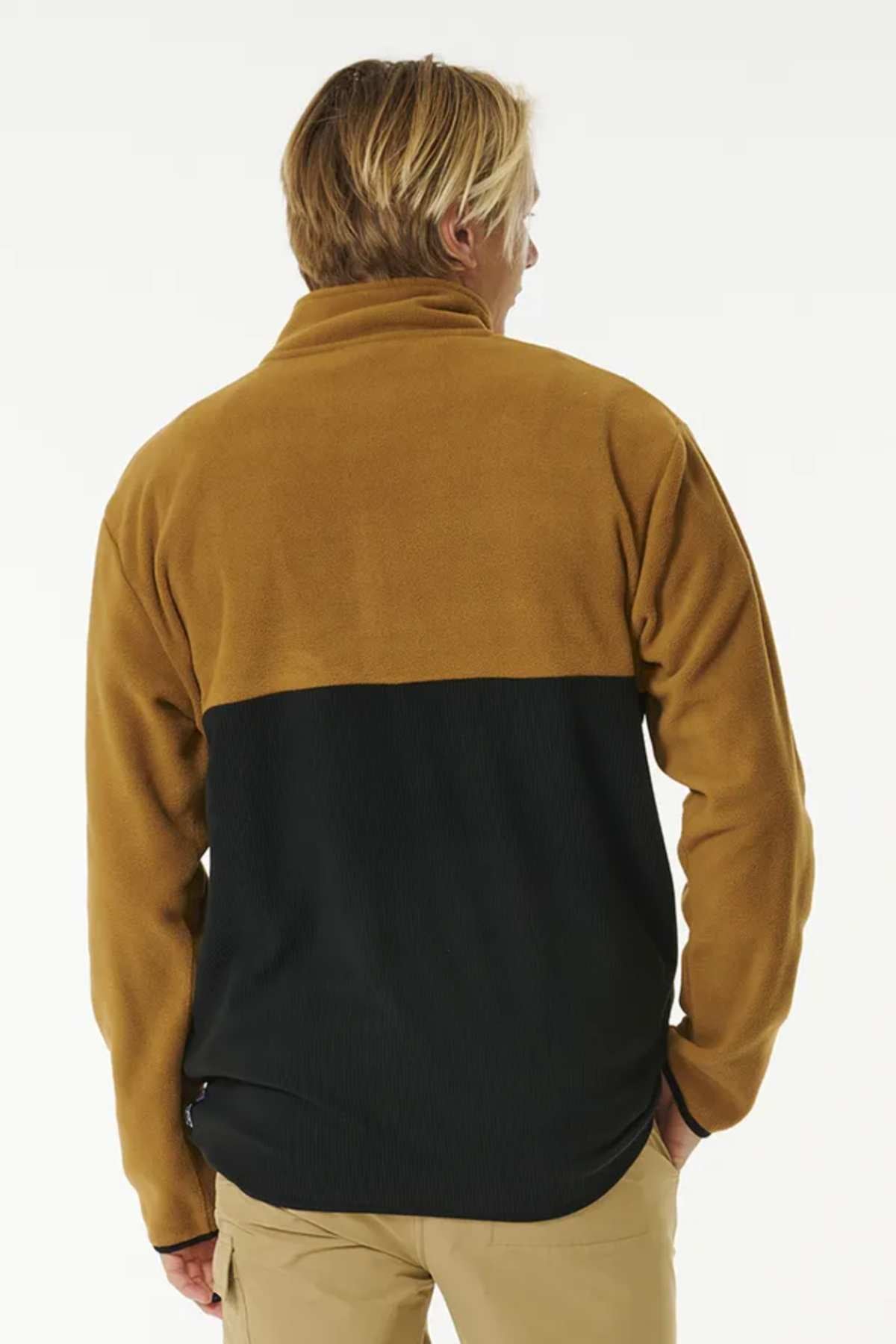 A person with blonde hair is shown from the back wearing a two-toned Rip Curl Journey Polar Fleece Crew. The top half of the jacket is camel brown, and the bottom half is black, featuring a chest pocket. The person is also wearing tan pants. The background is plain white.
