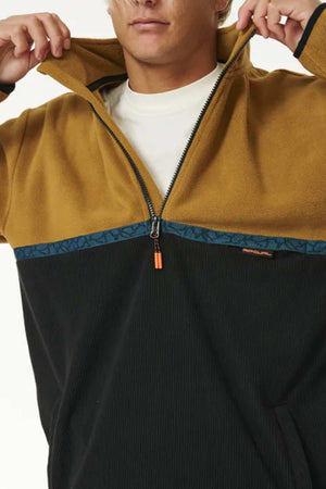 A person wearing a two-toned Rip Curl Journey Polar Fleece Crew with a mustard-yellow collar, upper chest, and sleeves, and a black lower section featuring a dark blue horizontal stripe near the half zip front. The person is partially zipping up the jacket, revealing a white undershirt beneath.