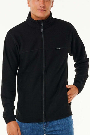 A person wearing a black Rip Curl Journeys Grid Polar Fleece with a full zip front and blue jeans poses with one hand on their hip against a plain, light background.
