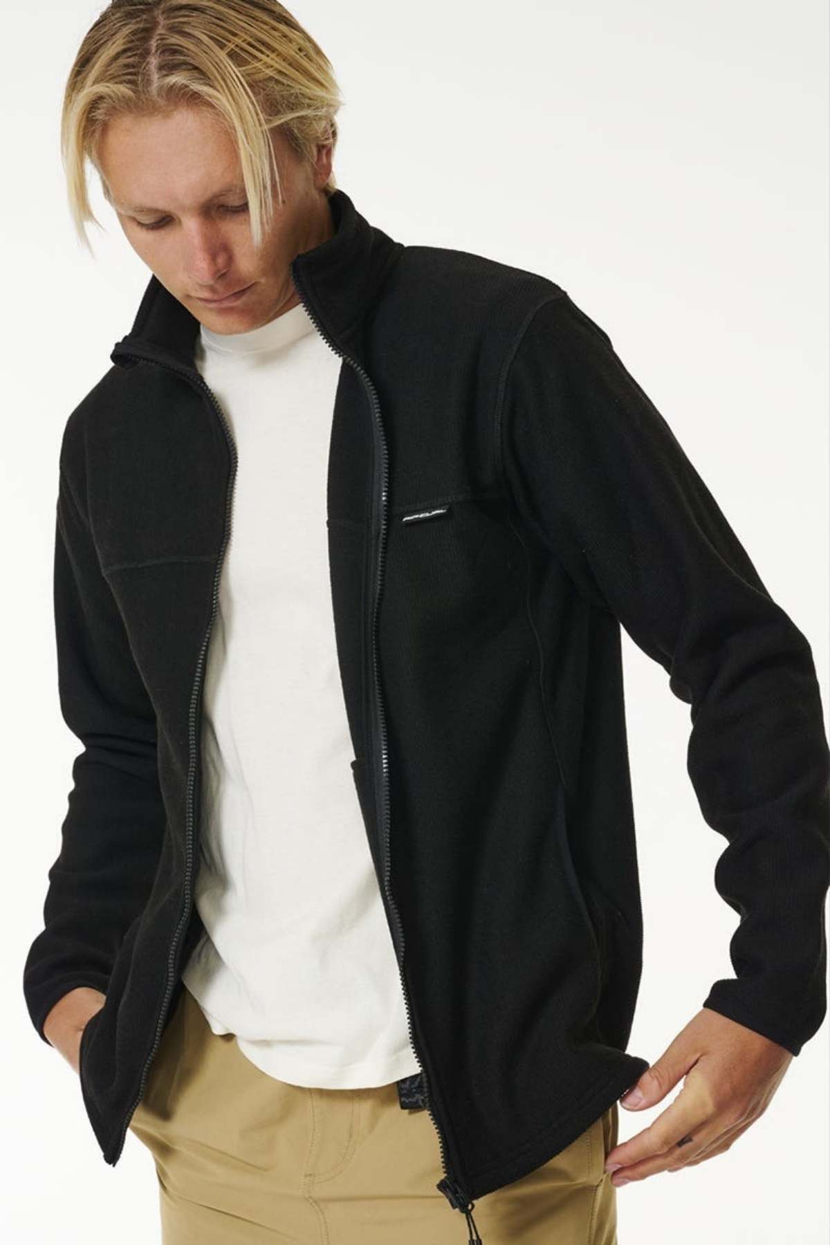 A person with blonde hair wears a Rip Curl Journeys Grid Polar Fleece black jacket in regular fit over a white shirt and beige pants, looking down while adjusting the full zip front, set against a plain light background.