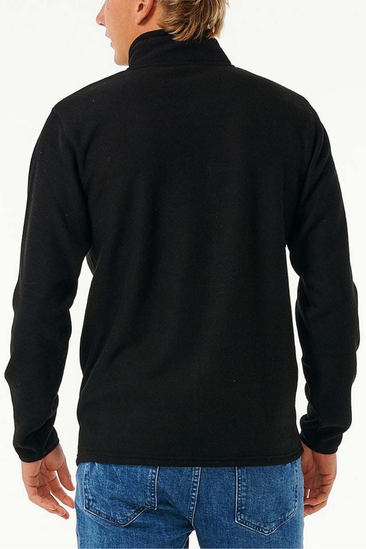 A person with short hair is seen from behind, wearing a black Rip Curl Journeys Grid Polar Fleece and blue jeans against a plain, light background.
