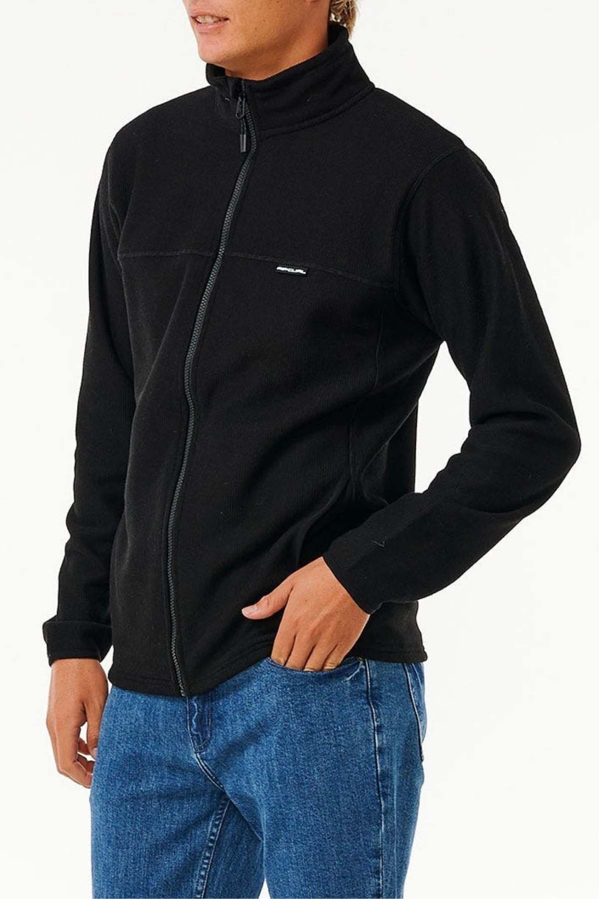 Wearing a Rip Curl Journeys Grid Polar Fleece black jacket with a full zip front and blue jeans, the person stands with one hand in a pocket against a plain white background, head turned so their face is not fully visible.