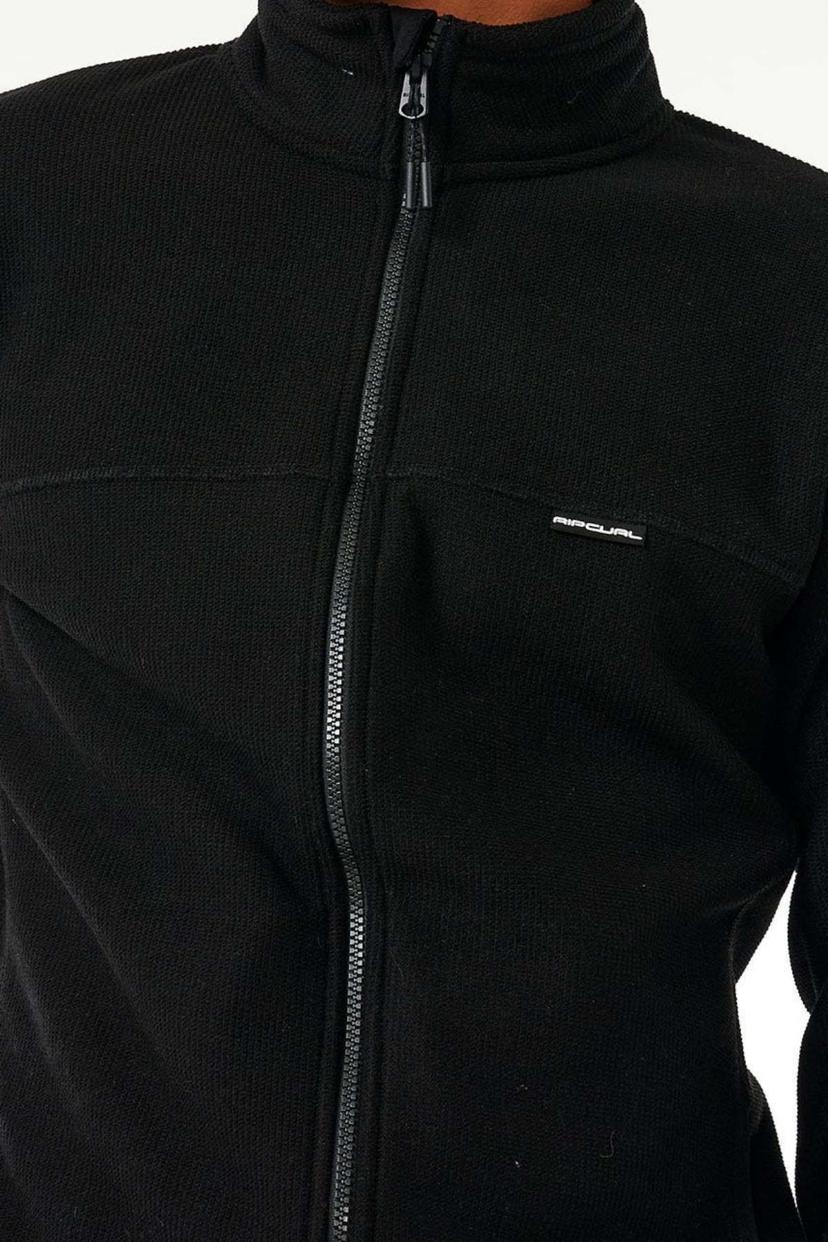 A person wears a black Rip Curl Journeys Grid Polar Fleece with a small logo on the left chest. The regular fit and full zip front are visible. Photo shows shoulders to waist against a plain light-colored background.