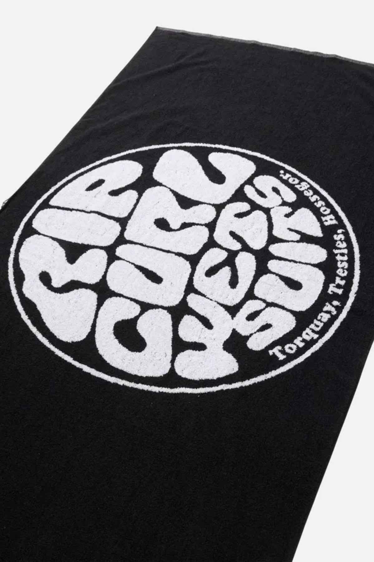 A striking design adorns the Rip Curl Logos Towel, featuring a bold black cotton fabric with a large circular white emblem displaying "Rip Curl, 50 Years of Surf" and "Torquay Australia" in stylized, bold lettering. The plain background enhances the iconic beach towel's distinctive look.