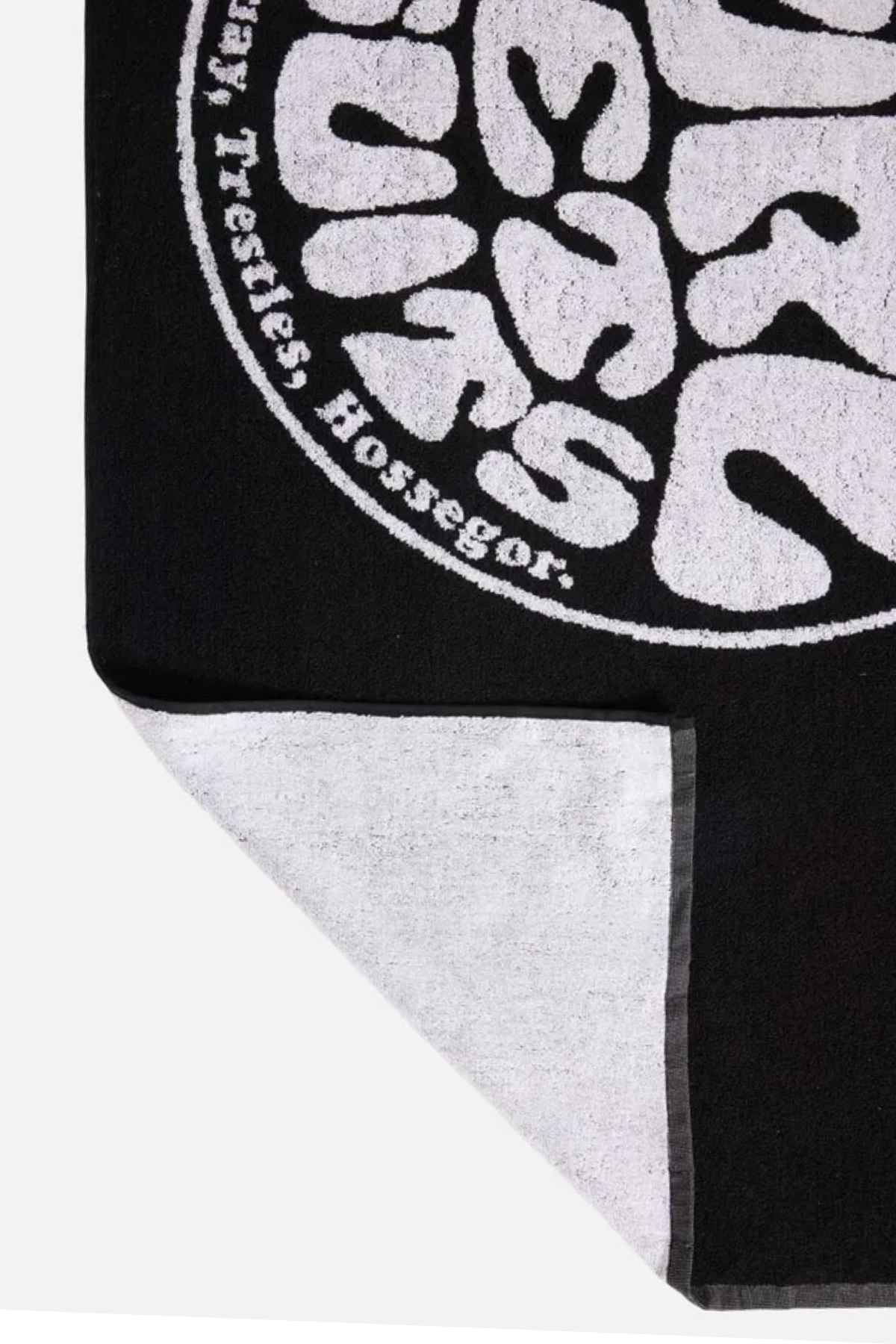 A Rip Curl Logos Towel in black showcases a white circular logo in the corner with stylized text and the words "Trestles, Hossegor." The corner is flipped over to reveal a lighter cotton underside.