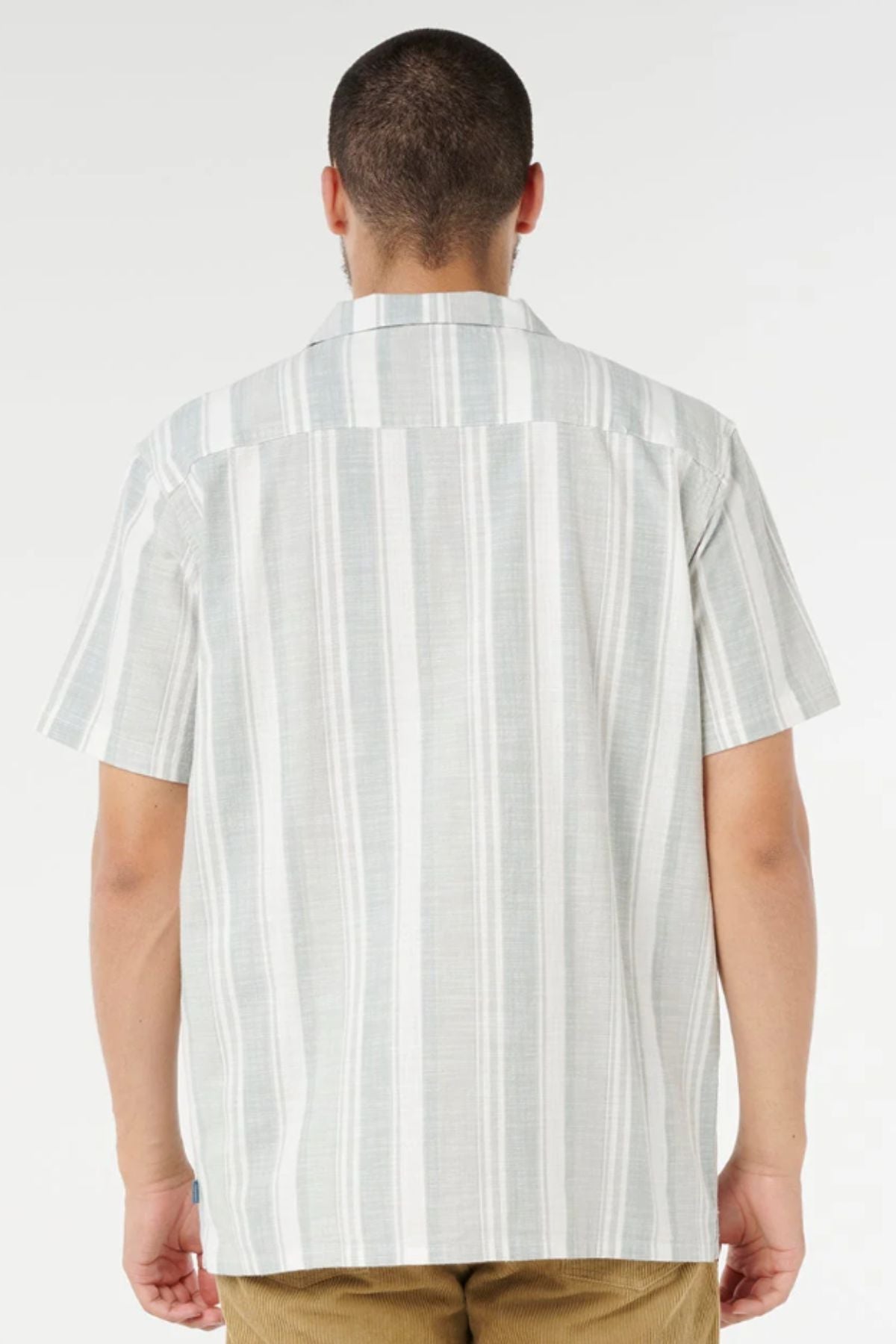 A person with short hair is shown from the back, wearing a Rip Curl Mod Cali Stripe Short Sleeve Shirt in light gray and white stripes, paired with light brown pants, standing against a plain white background.