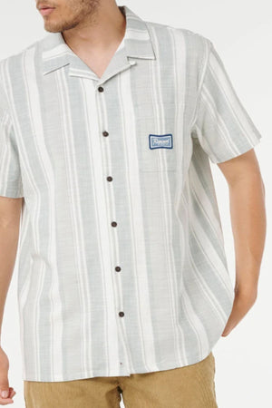 A man wearing the Rip Curl Mod Cali Stripe Short Sleeve Shirt with a small blue chest patch and tan corduroy pants stands with one hand in his pocket.
