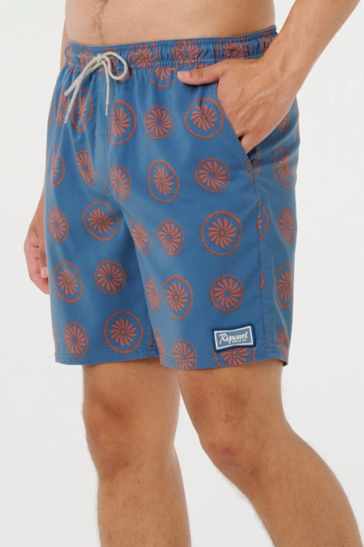 Wearing the Rip Curl Mod Cali Volley Short—blue with an orange circular pattern and drawstring waist—a person stands with one hand in a pocket. Quick-dry fabric and a Ripcurl logo patch finish the look.