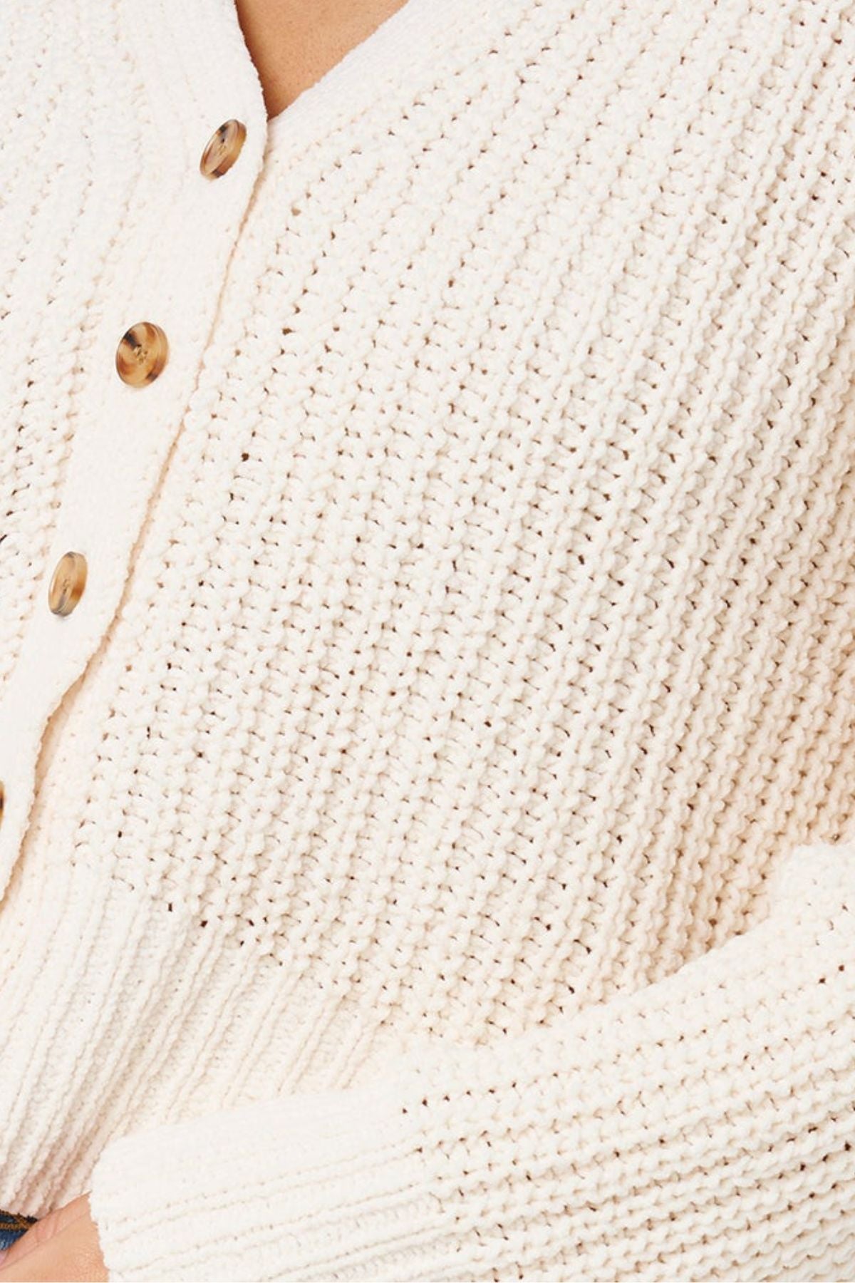 Close-up of the Rip Curl Neon Isles Cardigan, cream-colored with a chunky textured knit, ribbed detailing, brown buttons, and part of a hand resting at the bottom.