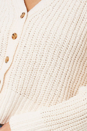 Close-up of the Rip Curl Neon Isles Cardigan, cream-colored with a chunky textured knit, ribbed detailing, brown buttons, and part of a hand resting at the bottom.