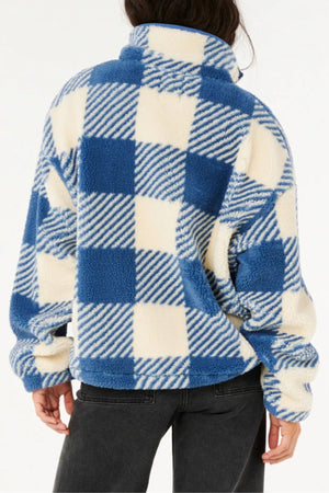 A person faces away from the camera wearing the Rip Curl Neon Isles Zip Through Polar Fleece in blue and white check, featuring a high collar and soft texture for a cozy, sustainable style, paired with dark pants.