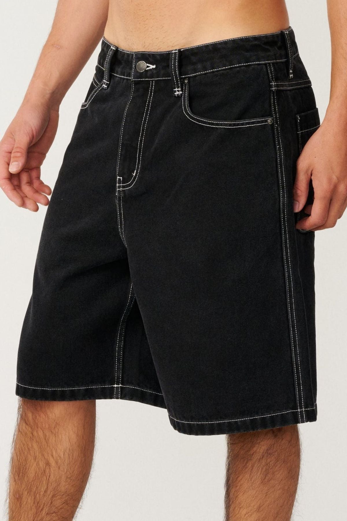 A person is shown from the waist to mid-thigh, wearing the Rip Curl Nu Dose Denim Walkshort in black with white stitching. These baggy fit shorts feature front and side pockets, a button closure, and evoke a classic skate style look.