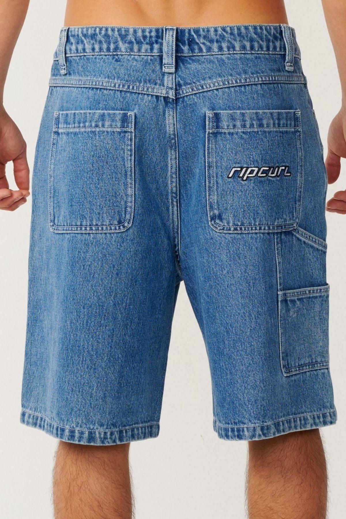 A person wearing Rip Curl Nu Dose Denim Walkshorts, featuring a baggy fit, large back pockets, and a "ripcurl" logo on one pocket, shown from the waist down and viewed from behind—a classic skate style look.