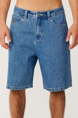 A person wearing the Rip Curl Nu Dose Denim Walkshort stands against a plain light background. The baggy fit shorts feature classic pockets, belt loops, and a visible button, with a small logo on the left pocket that channels subtle skate style. Only the torso and upper legs are visible.