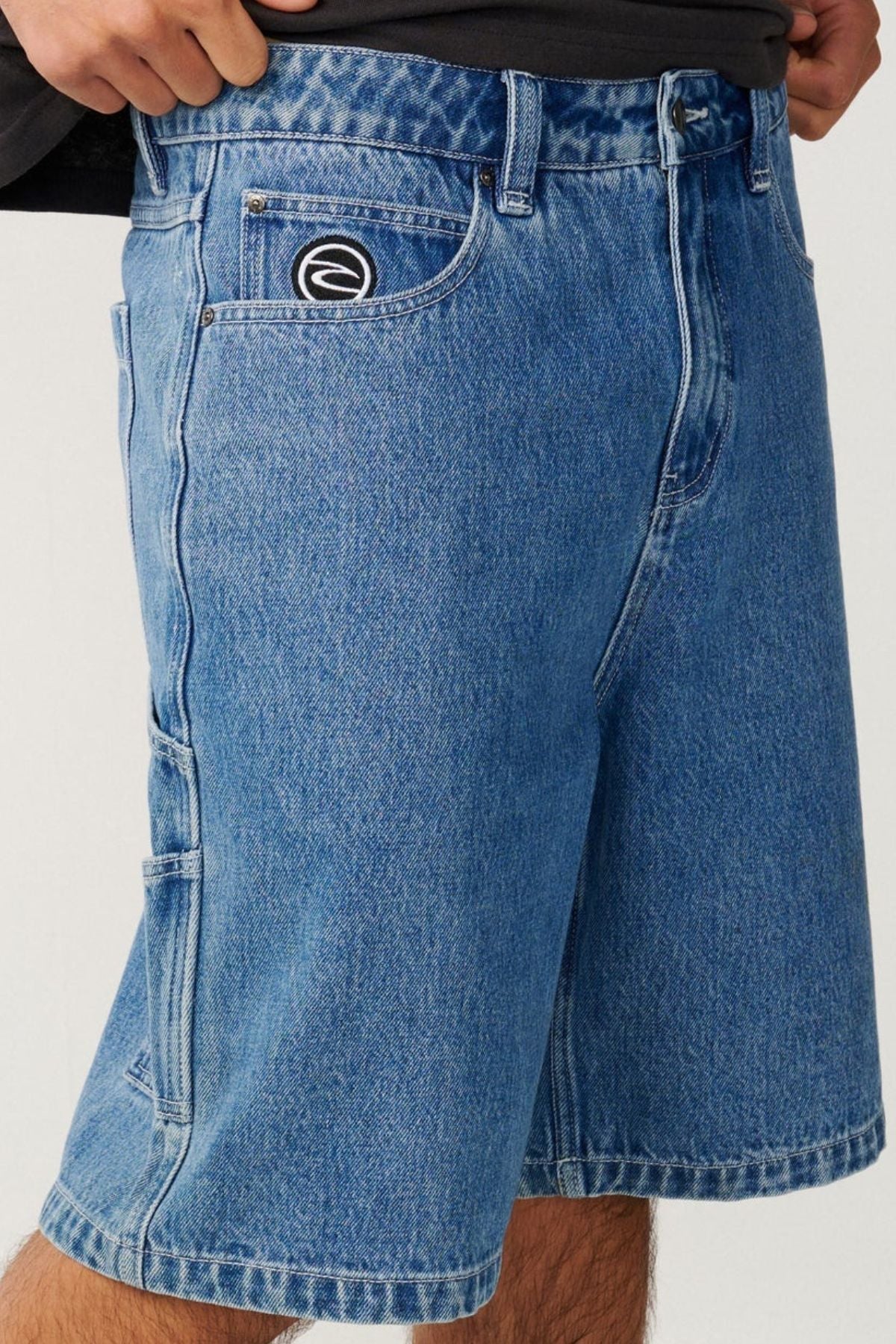 A person wearing the Rip Curl Nu Dose Denim Walkshort in knee-length blue denim, featuring a black and white circular logo patch on the front pocket, styled in a classic skate look with a black shirt tucked at the waist.