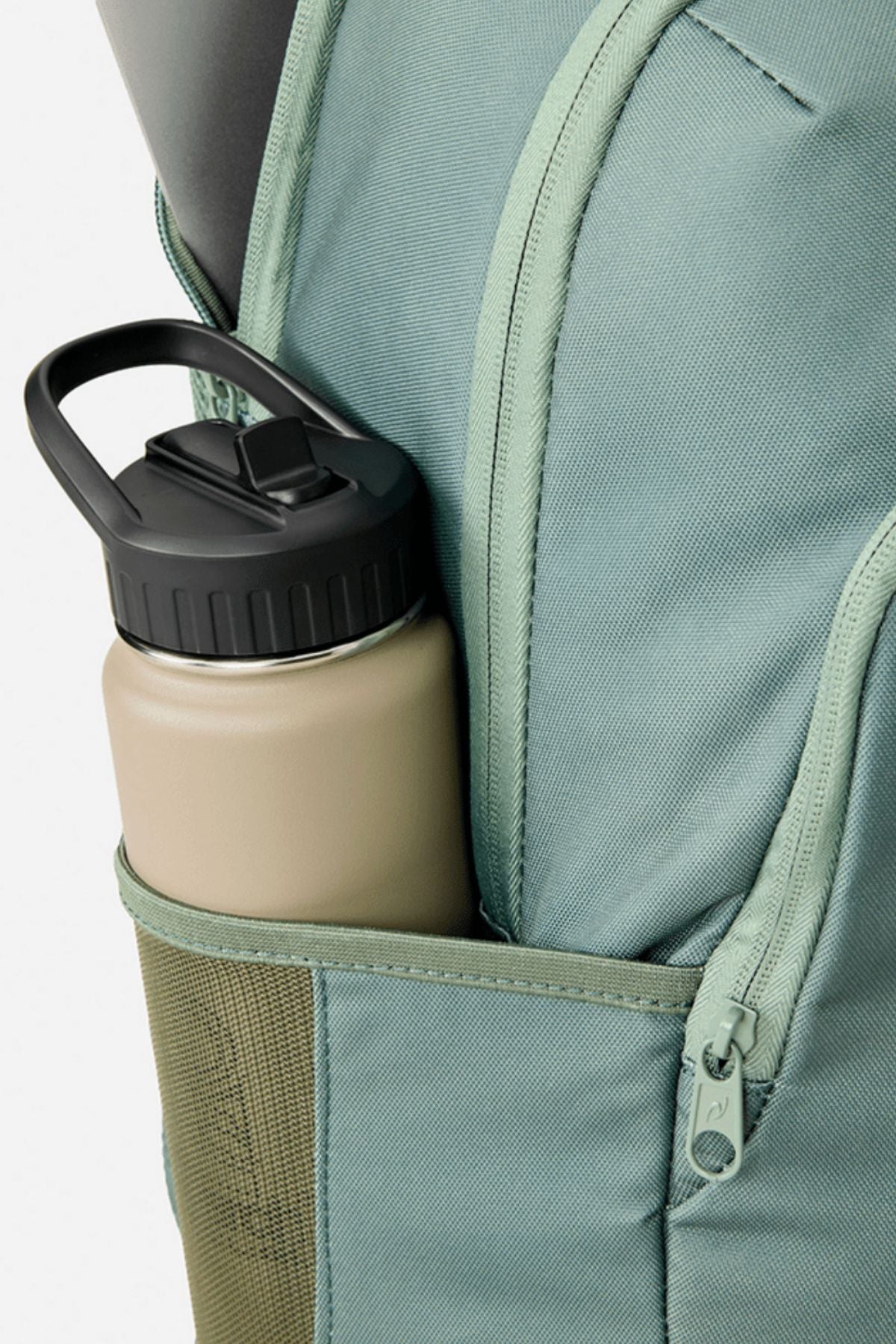 A beige reusable water bottle with a black lid sits in the side pocket of a light green Rip Curl Ozone 2.0 30L Backpack.
