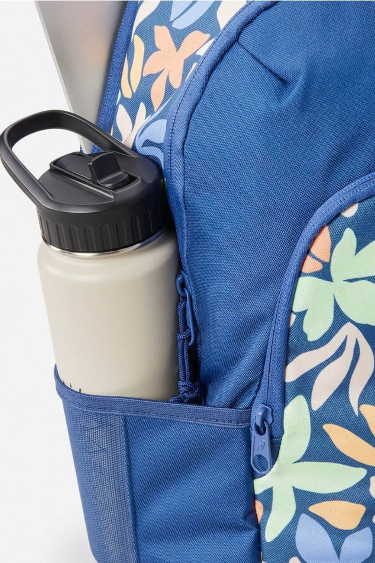 A close-up of the Rip Curl Ozone 2.0 30L Backpack in blue floral print, showing a zippered pocket, laptop compartment, and a beige reusable water bottle in the mesh side pocket.