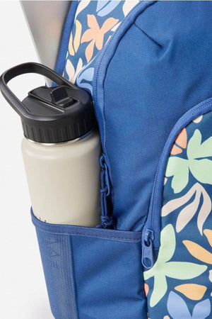 A close-up of the Rip Curl Ozone 2.0 30L Backpack in blue floral print, showing a zippered pocket, laptop compartment, and a beige reusable water bottle in the mesh side pocket.