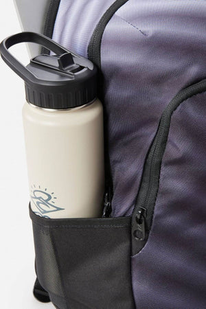 A white water bottle with a black lid rests in the side mesh pocket of a Rip Curl Ozone 30L Faded Slant Backpack. The bottle has a sketched design, and the mesh pocket matches the backpack's breathable straps and convenient laptop section.