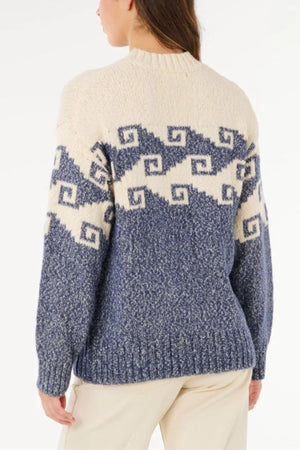 A person with long brown hair wears light-colored pants and the Rip Curl Pacific Coast Sweater in cream and blue, featuring a geometric zigzag pattern, standing with their back to the camera to display its coastal style.