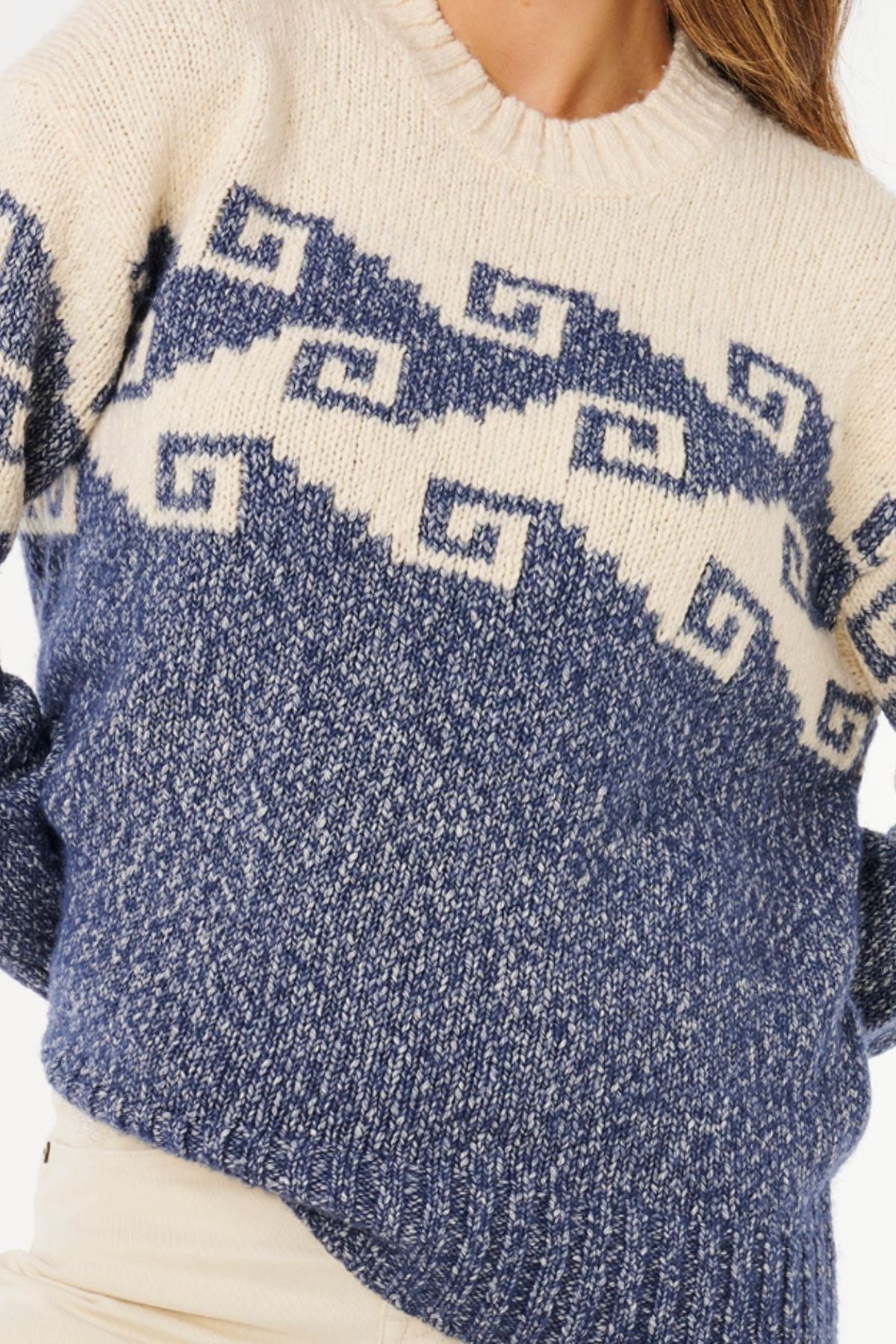 A person wearing the Rip Curl Pacific Coast Sweater in cream and navy blue with a geometric jacquard design, paired with light pants. Only the torso and part of the head are visible.