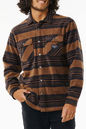 A person is wearing a brown and navy blue striped Rip Curl Party Pack Polar Fleece Shirt with chest pockets, standing with one hand in their pocket. The shirt has a casual, warm appearance, suitable for cooler weather. Made from recycled polyester, it's perfect for both style and sustainability. The person's face is partially visible, displaying a smile.