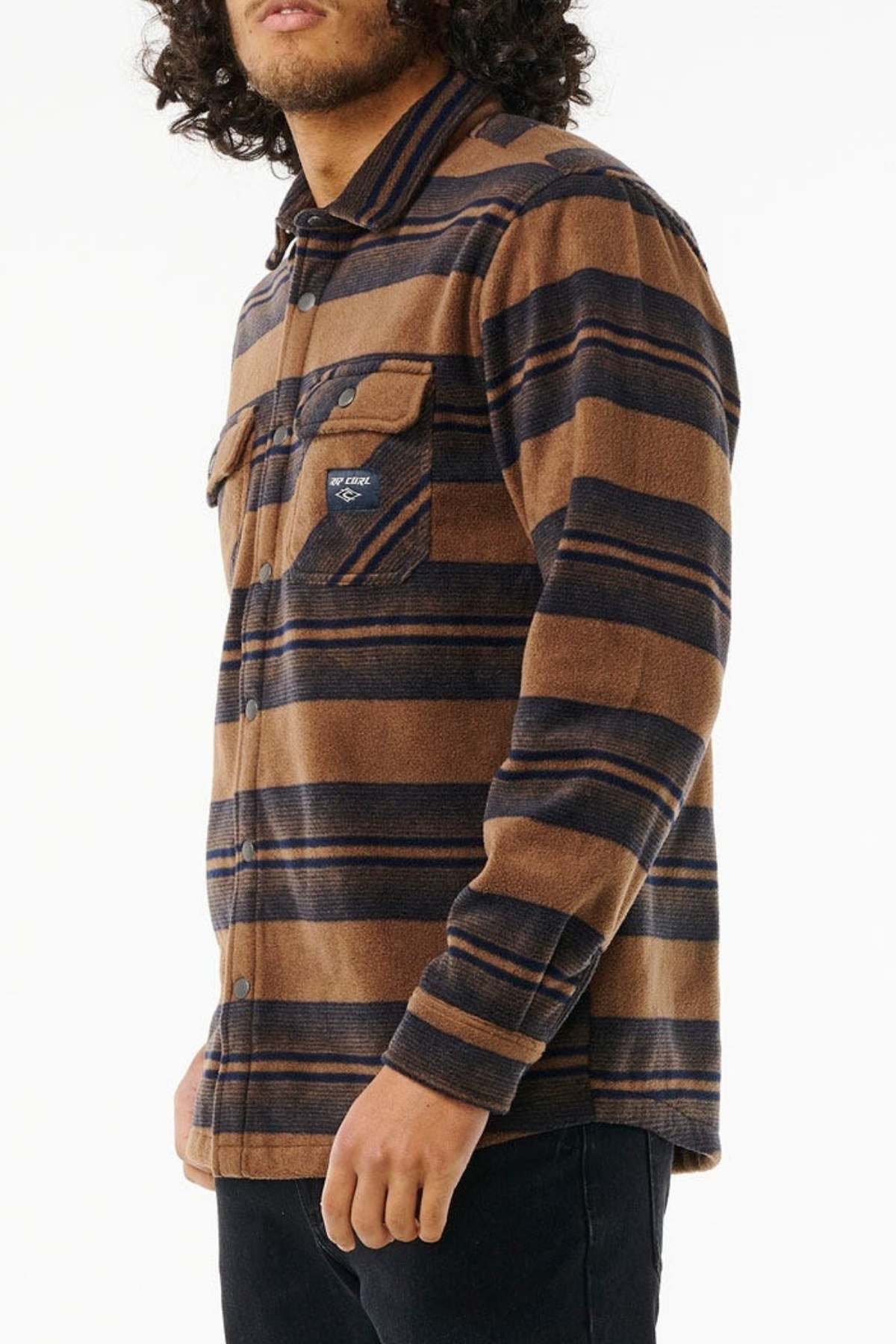 A person with curly hair wearing a brown and black striped Rip Curl Party Pack Polar Fleece Shirt, made from recycled polyester, stands against a white background facing slightly to the left. One hand is visible resting by their side while the other is partially obscured—perfect for cooler weather.