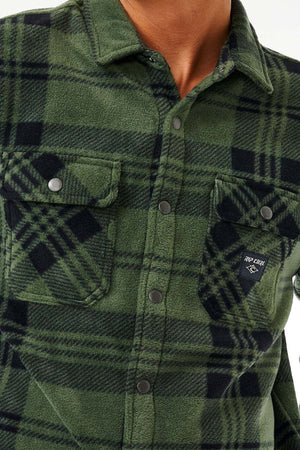 Close-up of a person wearing the Rip Curl Party Pack Polar Fleece Shirt in green and black plaid, an ideal choice for cooler weather. This shirt is designed with metal snap buttons running down the front and features two front pockets, each secured with a flap and button. One pocket displays a small patch with a partially visible logo and text.