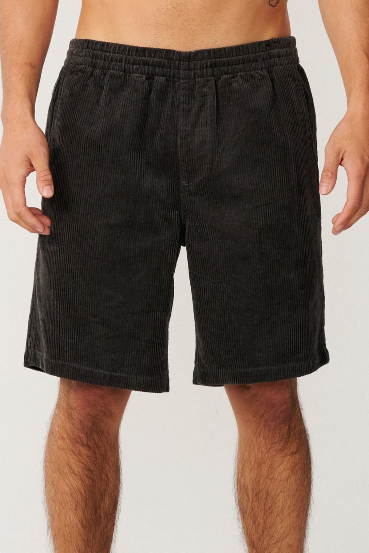A person wearing Rip Curl Premium Surf Cord 19" Volley black corduroy shorts stands against a plain light background, with only their lower torso and upper legs visible.