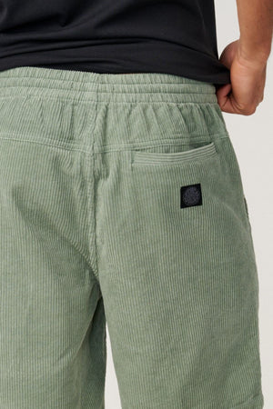 Close-up of a person wearing the Rip Curl Premium Surf Cord 19" Volley in light green, featuring corduroy fabric, an elastic waistband, and a back pocket for relaxed comfort reminiscent of your favorite cotton shorts. The person pairs the shorts with a black shirt and rests their hand on their hip.