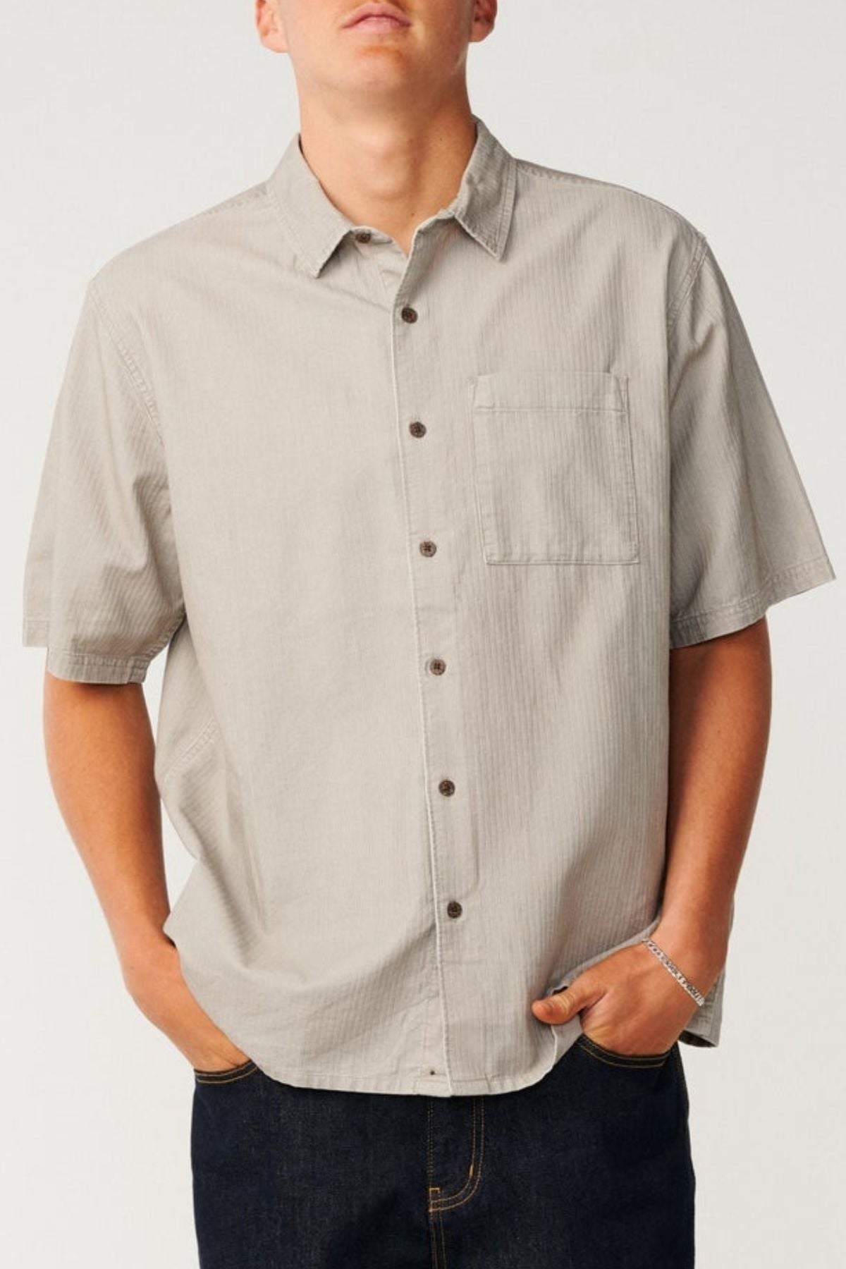 A person wearing a light gray Rip Curl Premium Surf Cord Short Sleeve Shirt featuring a chest pocket, paired with dark jeans and standing with hands in pockets against a plain background—a perfect example of casual wear with a touch of vintage aesthetic.