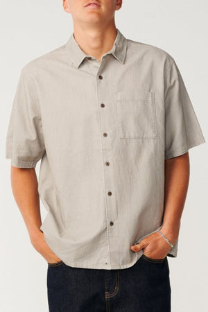 A person wearing a light gray Rip Curl Premium Surf Cord Short Sleeve Shirt featuring a chest pocket, paired with dark jeans and standing with hands in pockets against a plain background—a perfect example of casual wear with a touch of vintage aesthetic.