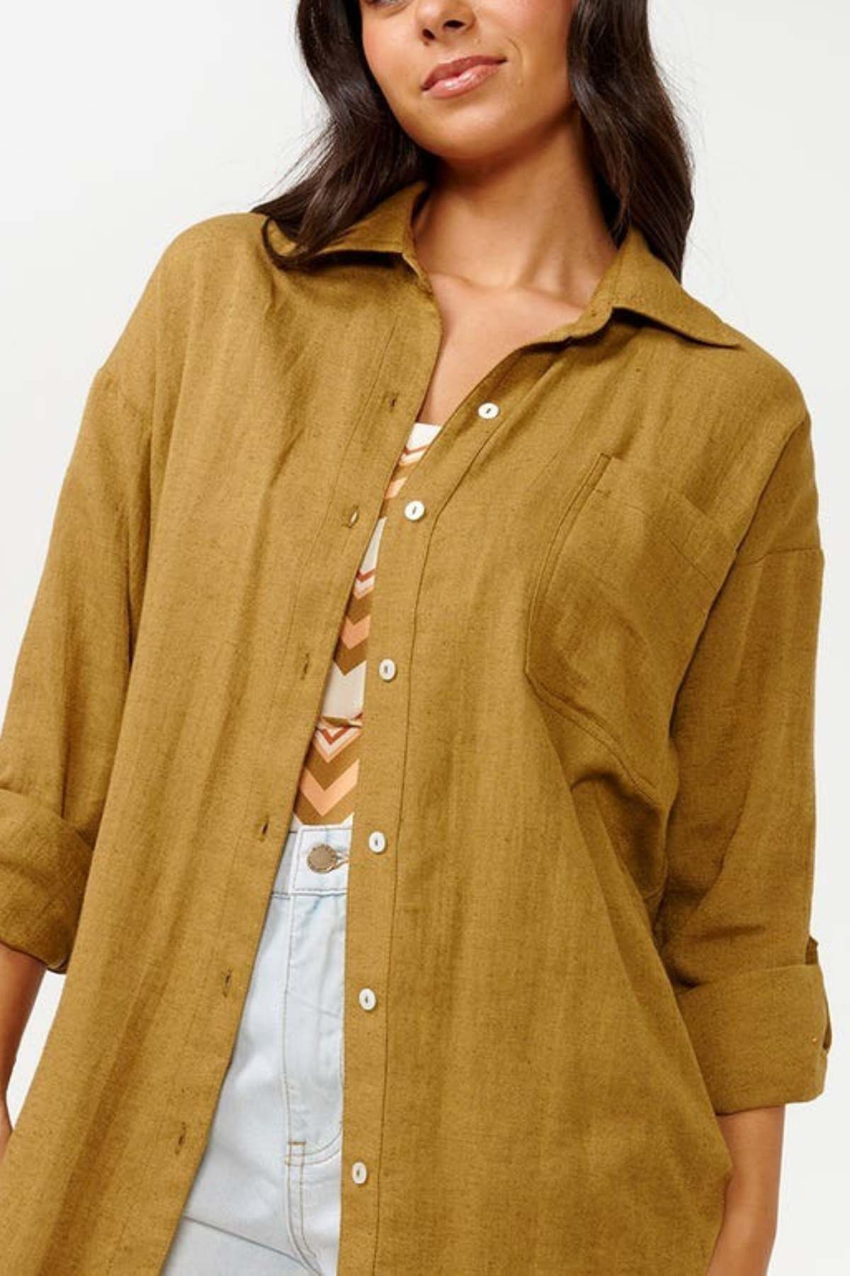 A woman dressed in a relaxed fit mustard-colored Rip Curl Premium Surf Holiday Shirt, layered over a patterned top. She has long, dark hair and sports light blue jeans. The shirt features cuffed sleeves and a chest pocket.