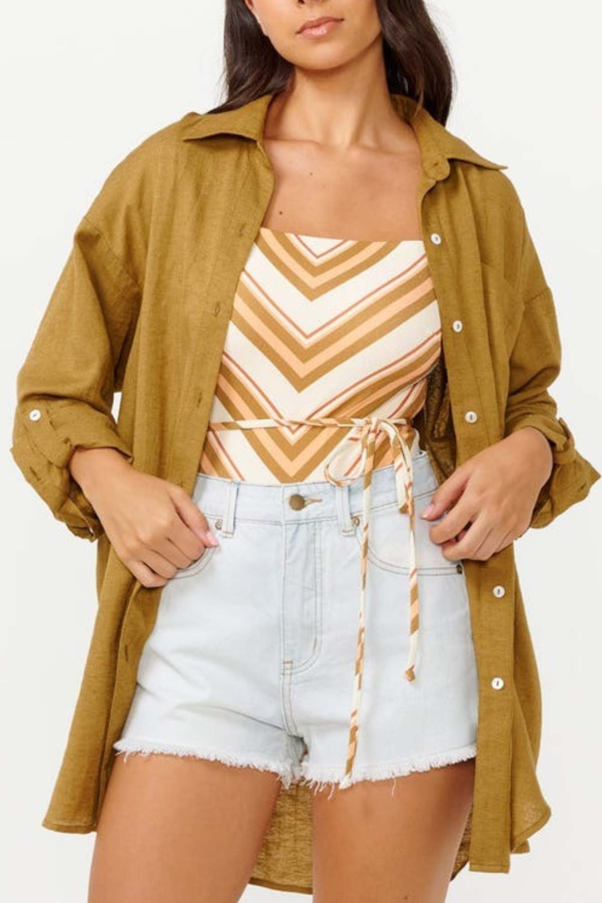 A woman personifies surf style with a relaxed-fit, mustard-colored Rip Curl Premium Surf Holiday Shirt layered over a striped sleeveless top tied at the waist. She finishes her look with light denim shorts, casually tucking her hands into the pockets.