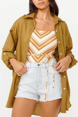 A woman personifies surf style with a relaxed-fit, mustard-colored Rip Curl Premium Surf Holiday Shirt layered over a striped sleeveless top tied at the waist. She finishes her look with light denim shorts, casually tucking her hands into the pockets.