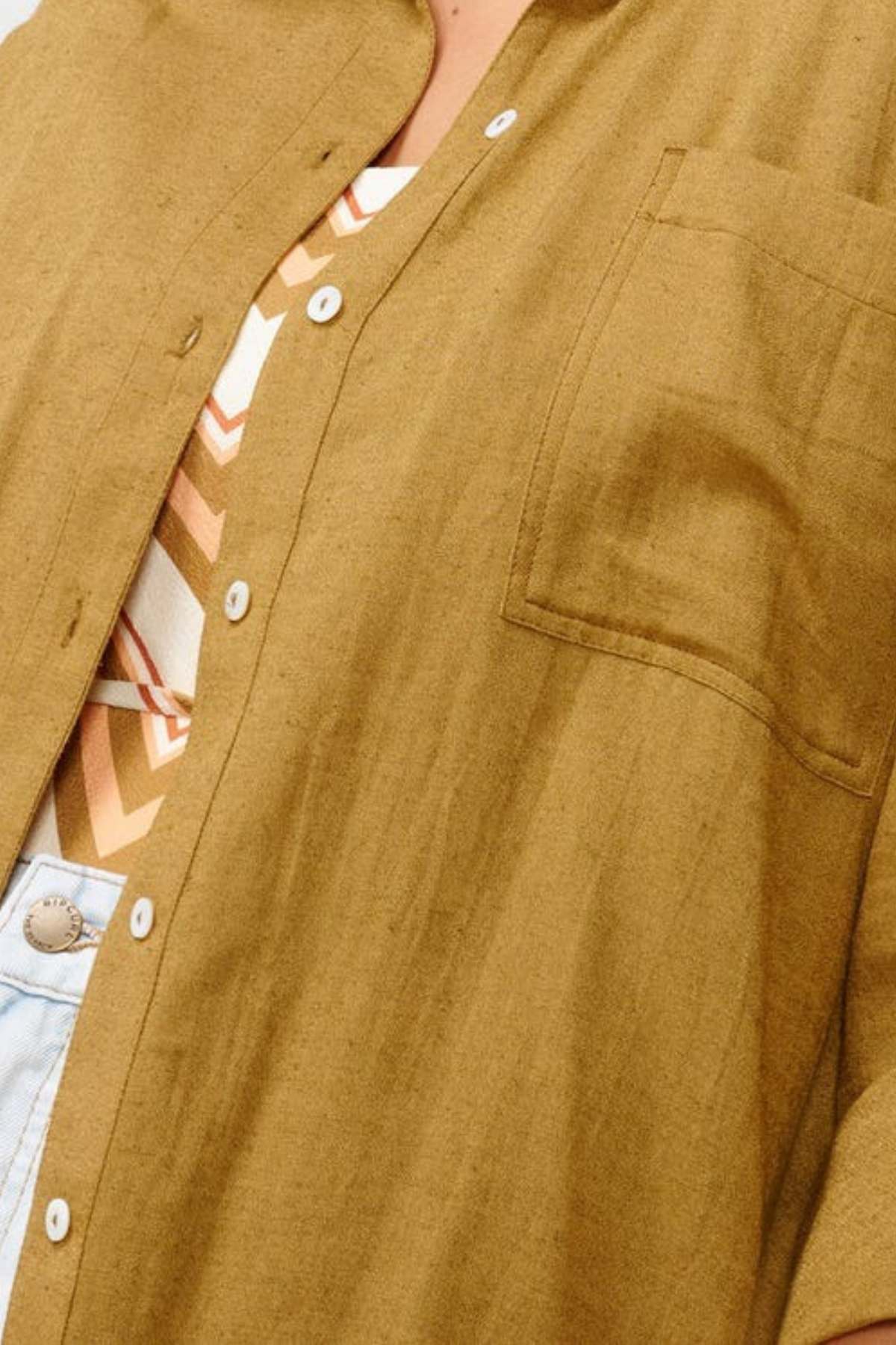 Close-up of a person wearing a relaxed fit Rip Curl Premium Surf Holiday Shirt in brown, showcasing its front pocket over an orange, white, and brown striped surf style top. The lower button is undone, revealing part of blue jeans.
