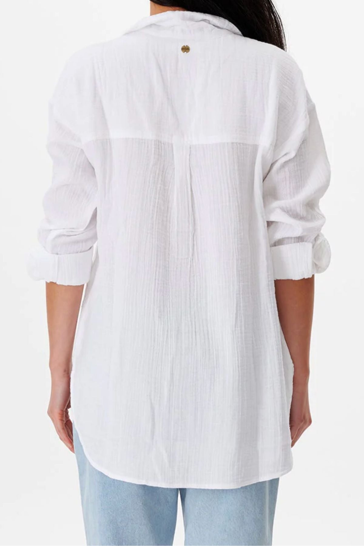 A person faces away, wearing the Rip Curl Premium Surf Long Sleeve Button Up Shirt in a relaxed fit with rolled-up sleeves, paired with light blue jeans. The white surf-style shirt extends below the hips for casual comfort.