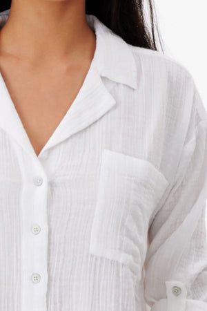 A close-up of a person wearing the Rip Curl Premium Surf Long Sleeve Button Up Shirt, featuring white textured cotton, a collar, chest pocket, white buttons, and a relaxed fit. Their dark hair is partially visible.