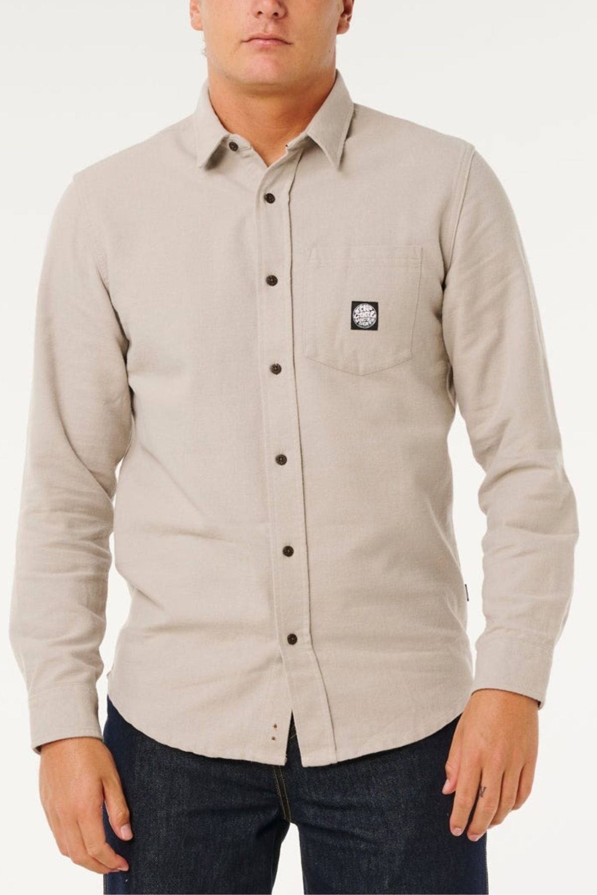 Wearing a light beige Rip Curl Premium Surf Flannel with a collar and black chest pocket patch, paired with dark blue jeans, a person stands against a plain light background, capturing a relaxed coastal lifestyle.