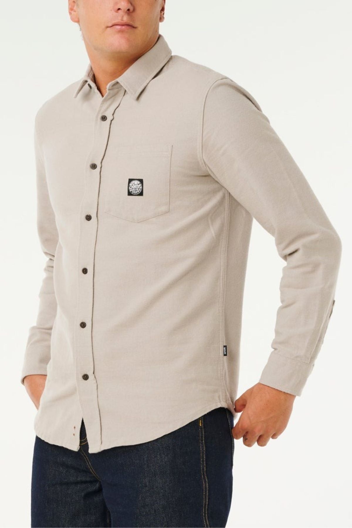 A person wearing a light beige Rip Curl Premium Surf Flannel with a chest pocket and dark buttons, paired with dark blue jeans, stands against a plain white background.