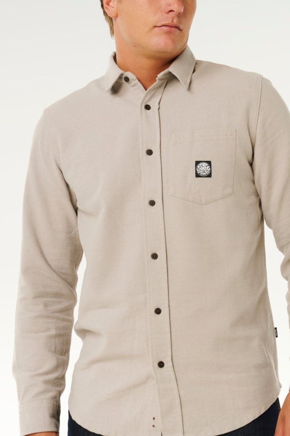 A man wearing a light beige Rip Curl Premium Surf Flannel with a black and white patch on the chest pocket, standing against a plain white background.