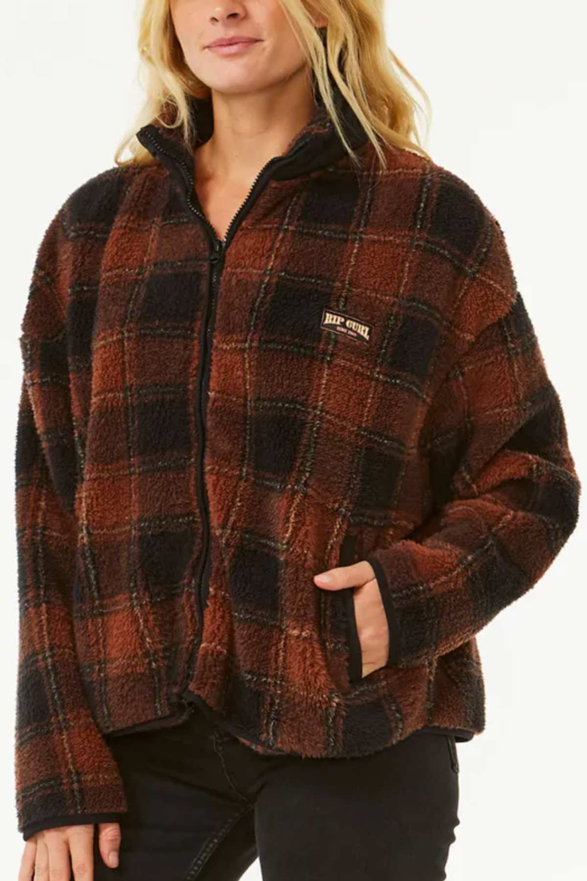 A woman wears the Rip Curl Sea Of Dreams Check Fleece, a stylish brown and black plaid zip-up jacket, with hands in pockets against a plain light background.