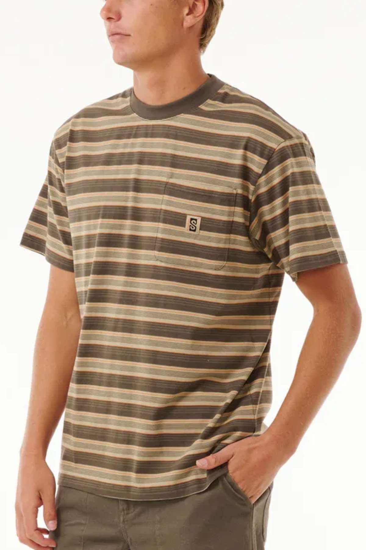 A person wearing a Rip Curl Search Stripe Tee made of 100% cotton in brown and beige, featuring a pocket logo, stands with their right hand in their pocket. They are photographed from the waist up against a plain background.