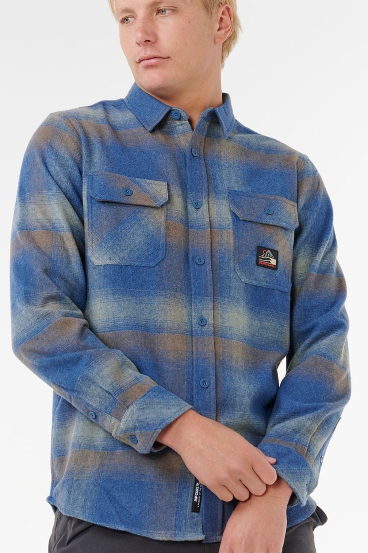 A person with blonde hair wears a blue and brown plaid Rip Curl Search Wool Flannel with two chest pockets, gazing to the side against a plain light background—ideal for effortless coastal living style.