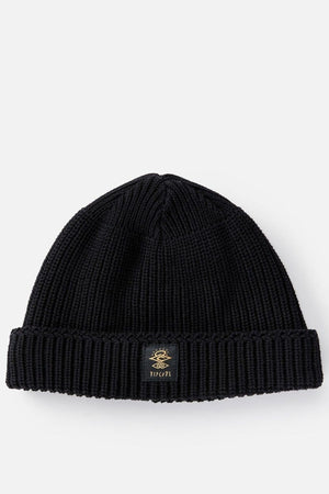 The Rip Curl Searchers Regular Beanie is a black, ribbed knit hat with a folded brim and a small gold-embroidered logo patch on the front center, shown laid flat on a light background.