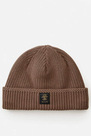The Rip Curl Searchers Regular Beanie is brown with a sherpa lining, folded brim, and features a small black patch with an embroidered design and "RIPOPI" in gold. It's displayed on a plain white background, ideal for those seeking warmth and style.