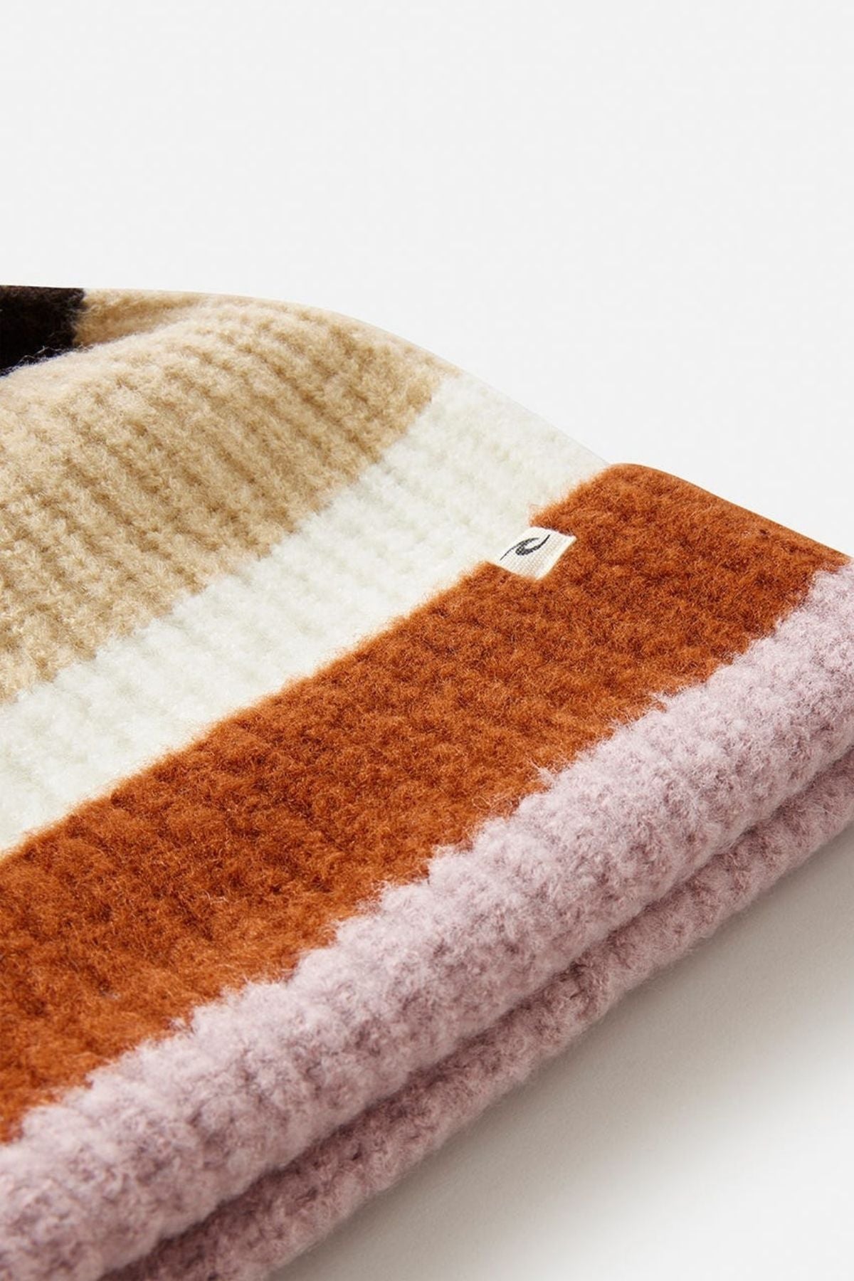 A close-up of the Rip Curl Sessions Regular Pom Pom Beanie in ribbed knit with wide tan, cream, rust orange, and light lavender stripes and a small fabric tag on the cream stripe—an essential winter accessory.
