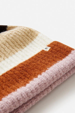 A close-up of the Rip Curl Sessions Regular Pom Pom Beanie in ribbed knit with wide tan, cream, rust orange, and light lavender stripes and a small fabric tag on the cream stripe—an essential winter accessory.