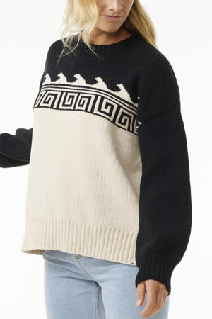 She wears the Rip Curl Soleil Knit Crew, an oversized color-block sweater with a black top featuring a white geometric horse pattern and cream recycled poly chenille bottom, paired with light blue jeans.