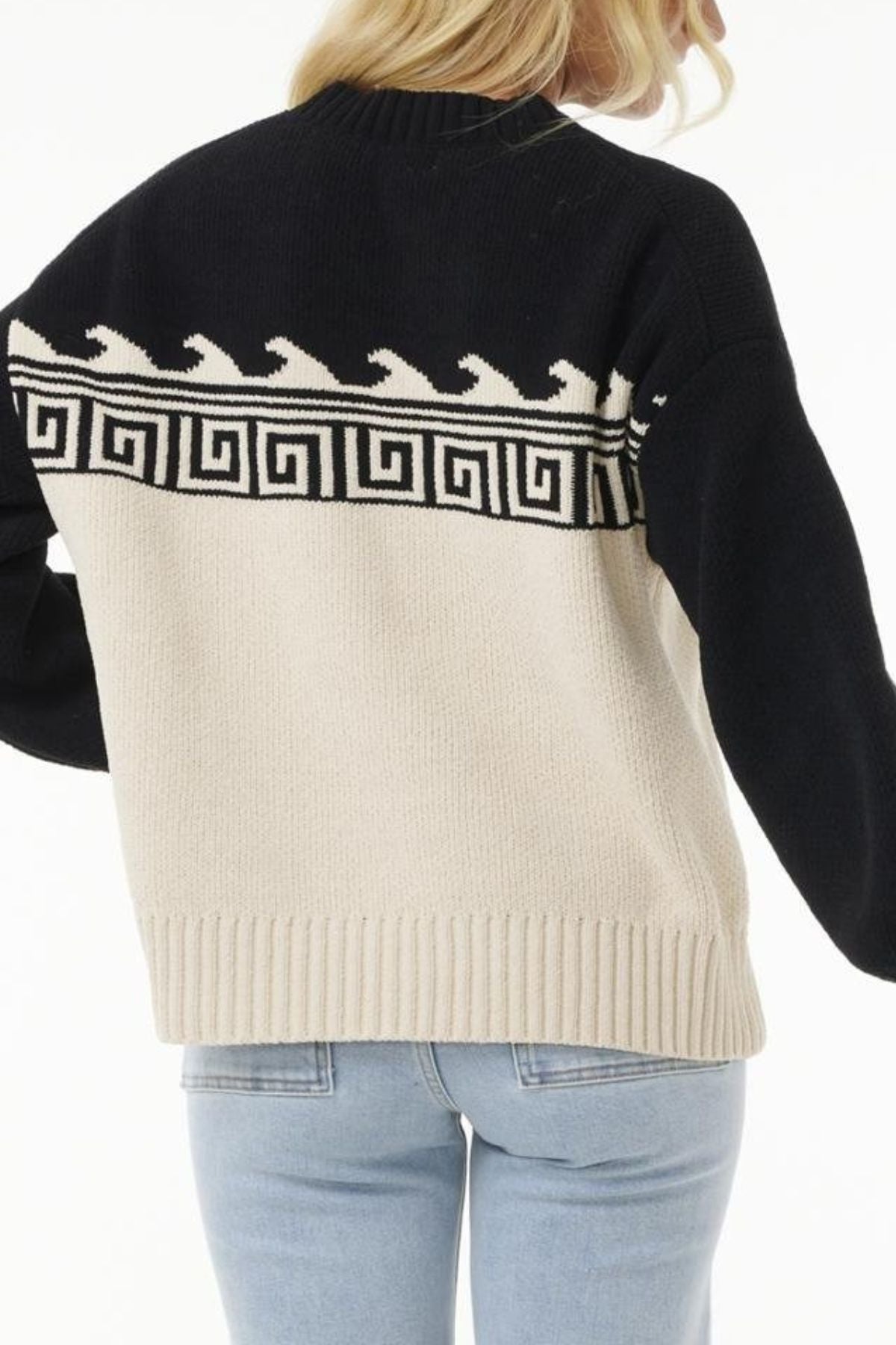 A person shown from behind wears light blue jeans and the Rip Curl Soleil Knit Crew, an oversized black and cream sweater featuring a Greek key and wave pattern across the back and shoulders.